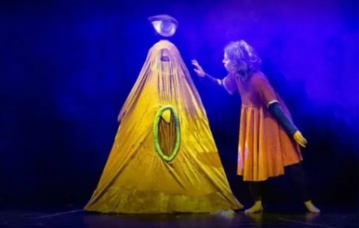 Escena de La ópera de los monstruos, con una figura amarilla en forma de cono y una actriz bajo luz azul.
