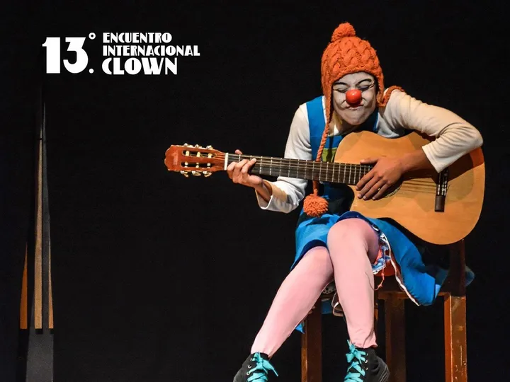 Una payasa con gorro naranja y nariz roja toca la guitarra sentada en un escenario oscuro.