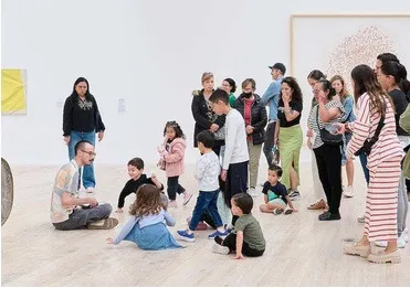 Un grupo de niñas, niños y adultos sentados en el piso de una sala blanca del museo, escuchando a una guía.