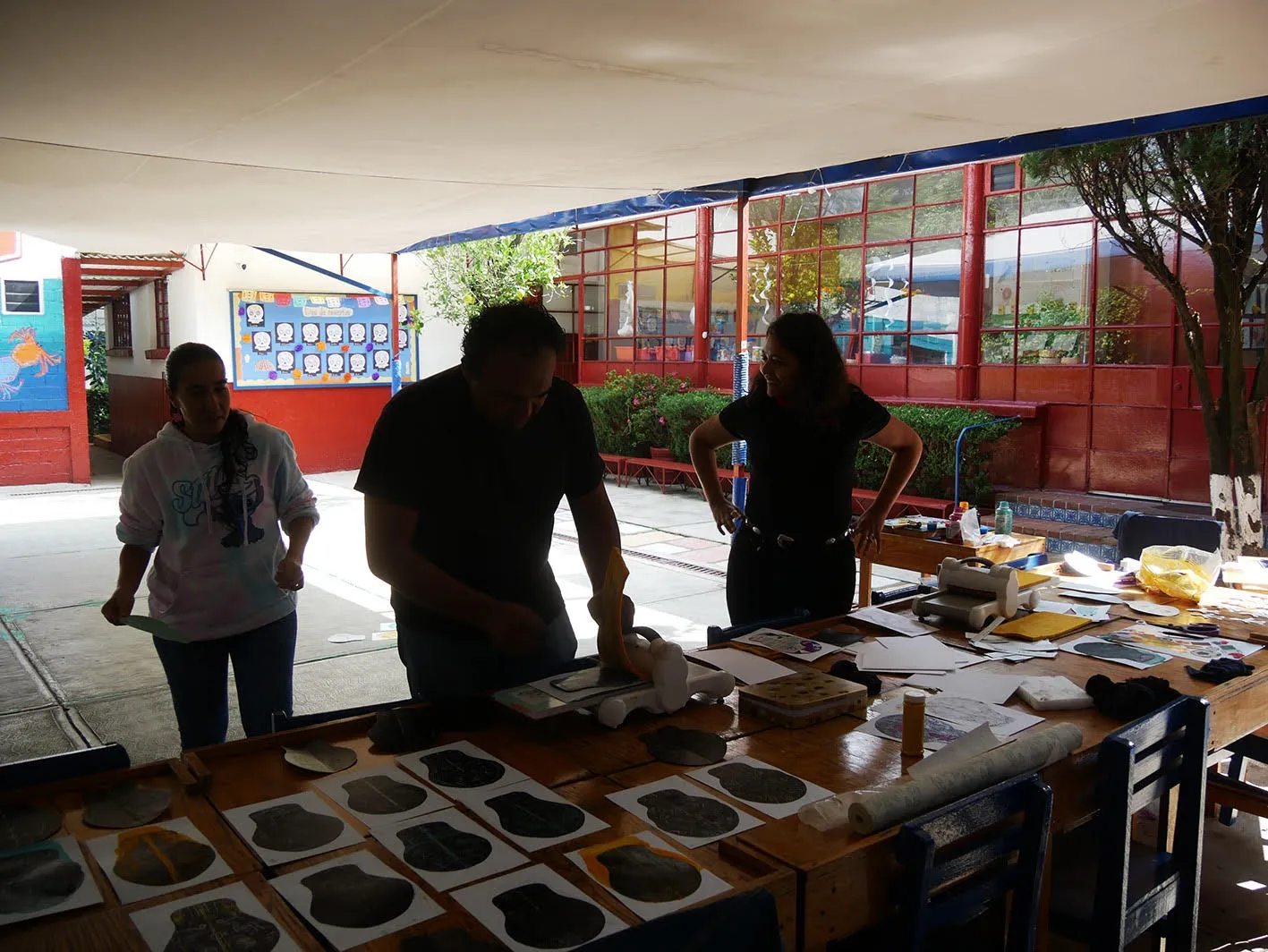 Tres personas trabajan alrededor de una mesa larga llena de impresiones, bajo un toldo en el patio.